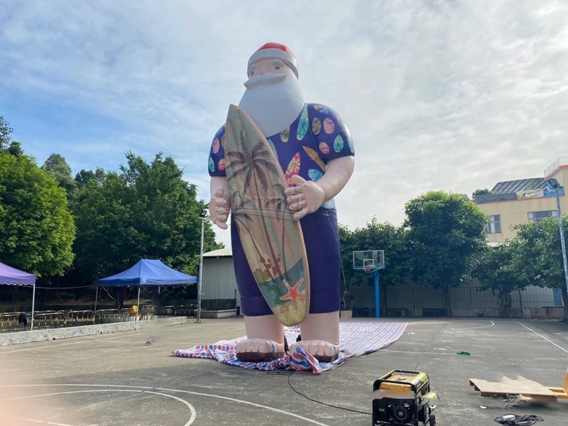 Giant inflatable Santa Claus surfing on the beach outdoors with a skateboard, beach themed Xmas inflatable decoration